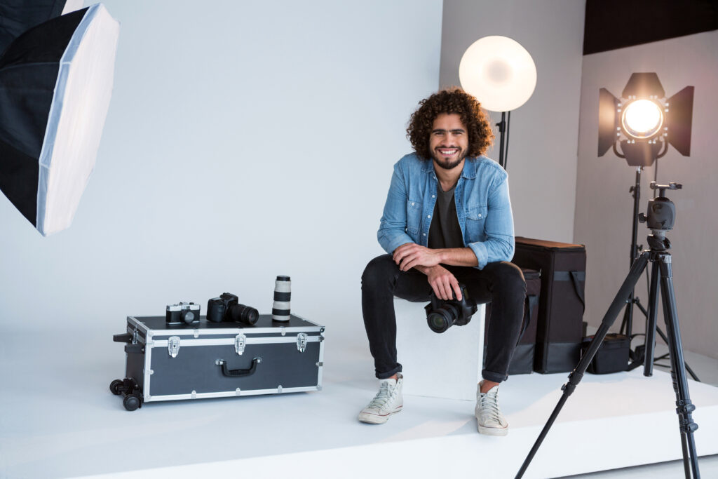 A smiling photographer sitting on a black box in his studio, holding a camera.