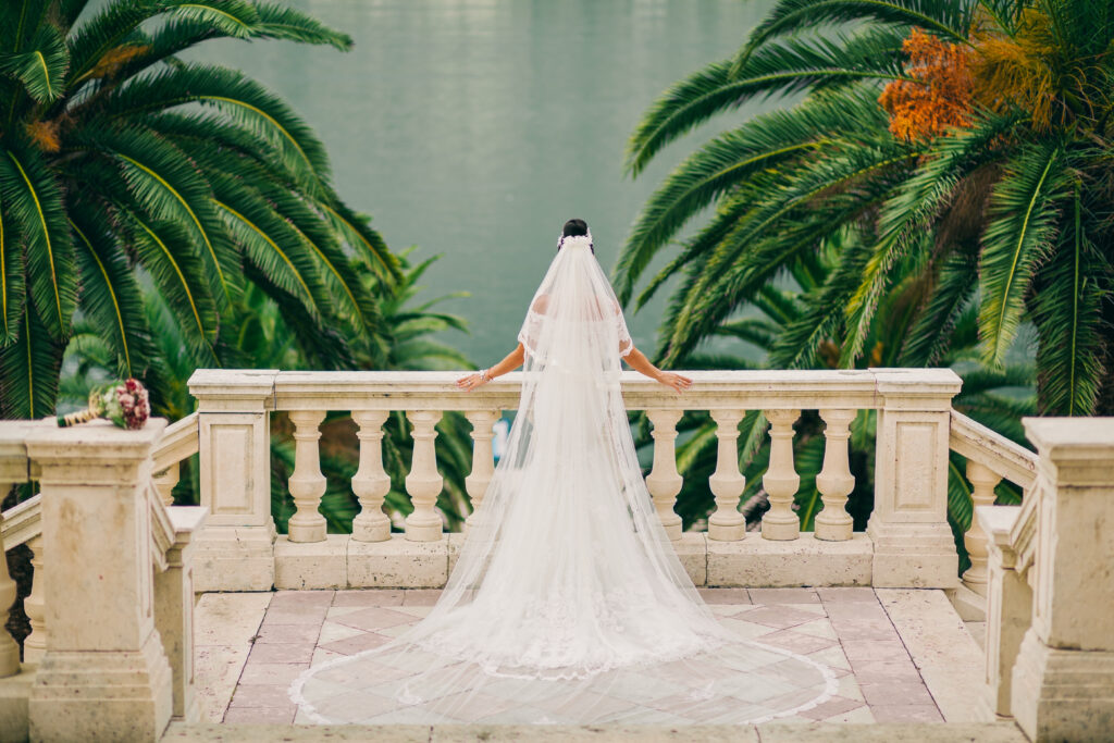 A bride in a wedding dress standing on a ledge, gazing at a serene lake flanked by two majestic palm trees.