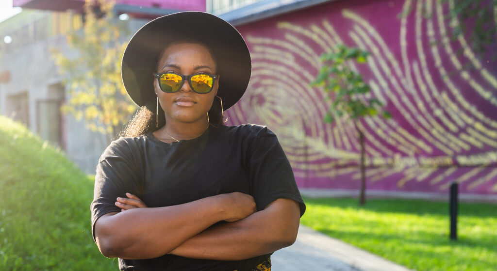 A woman wearing hats and sunglasses, crossing her arms in front of vibrant street art at Wynwood Walls.